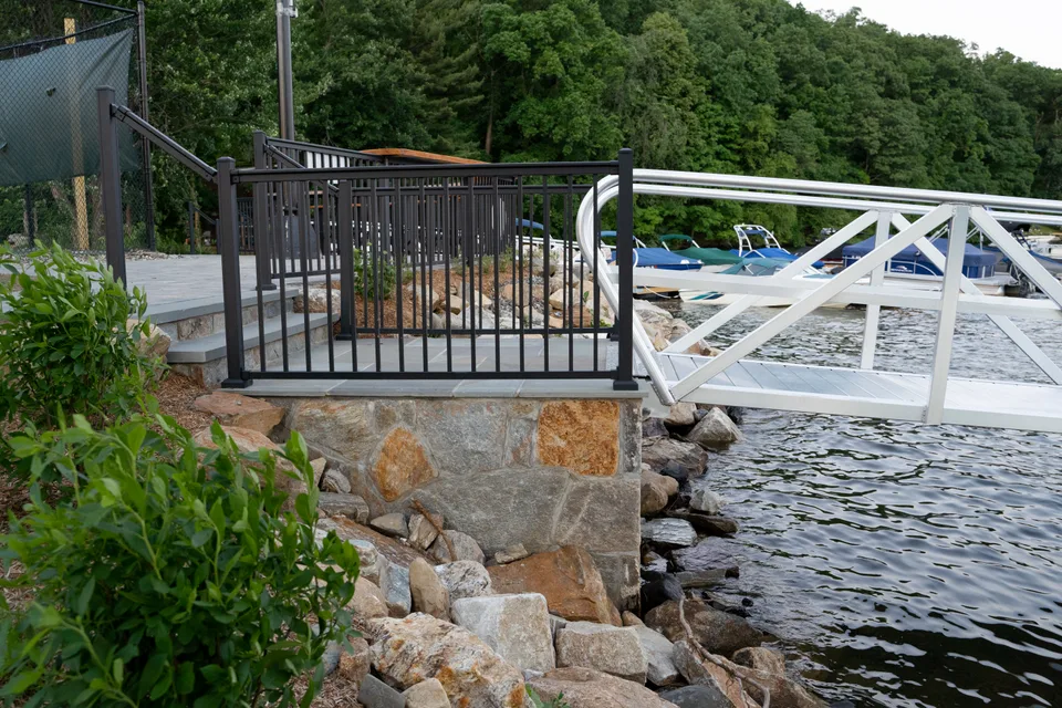 Fieldstone wall and steps with a black railing above the boulder shoreline at the dock — Exterior Services LLC
